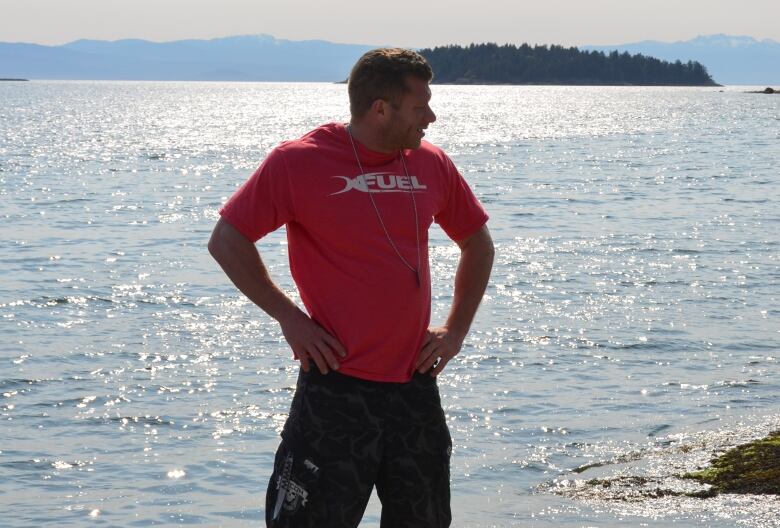 A young white man wearing a red T-shirt stands with his hands on his hips at the beach with the ocean behind him.