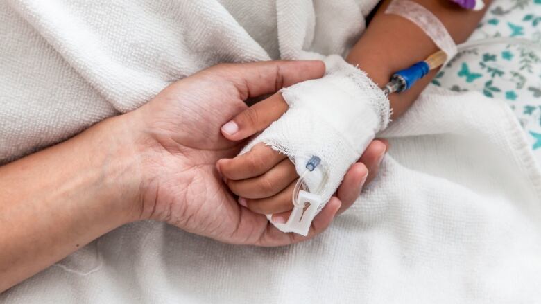 An adult holds a child's hand that has an IV in it. The hands are against a white blanket.