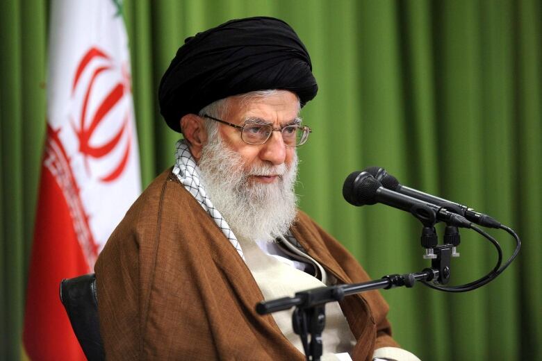 An old man in a turban sits in front of an Iranian flag.