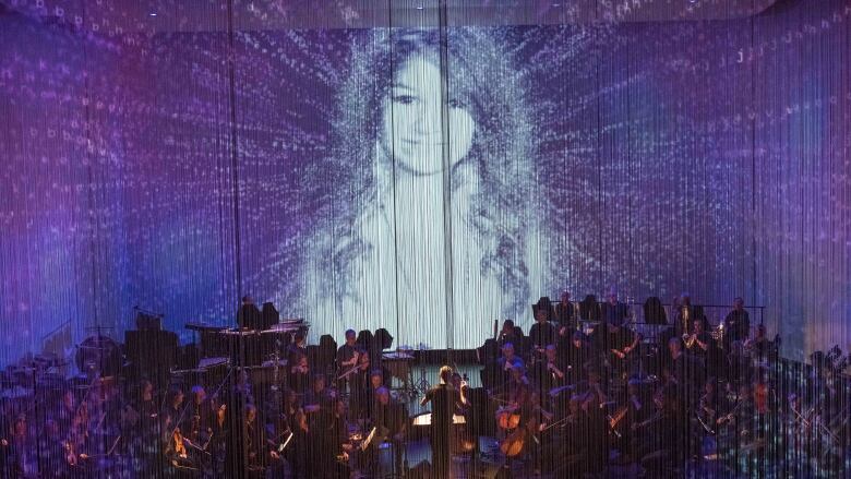 An orchestra performs while an ethereal image of Amanda Todd floats above the stage, surrounded by blue.