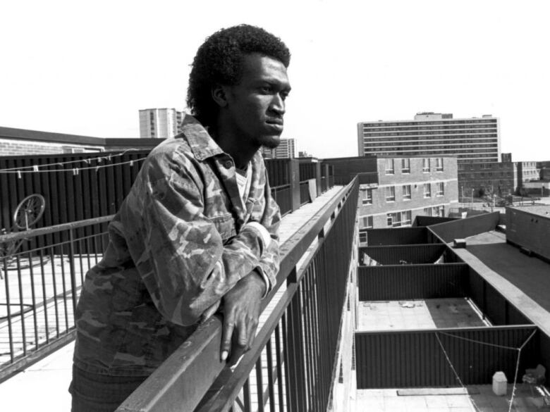 Still frame from the documentary Home Feeling: Struggle for a Community. Black-and-white shot of a Black man in a camo jacket leaning over a railing.