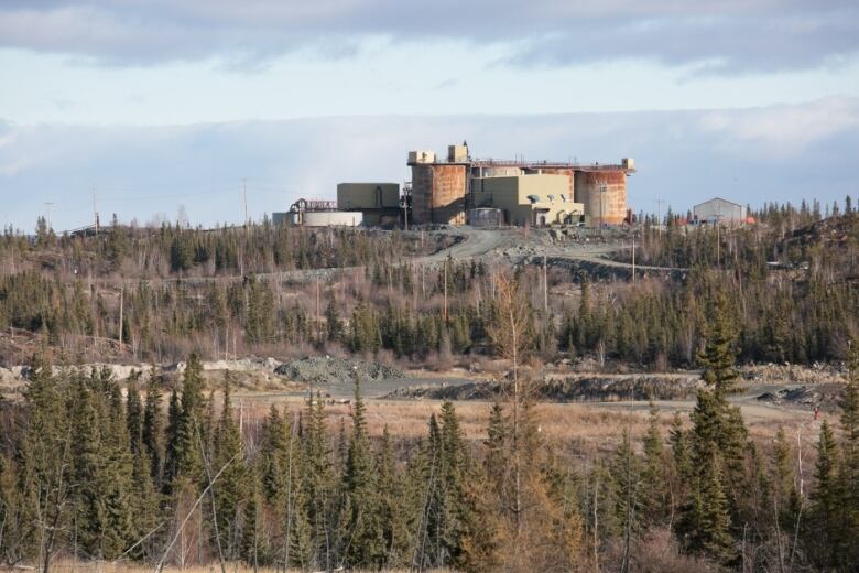 A building with huge round stack on the ends sits on top of a hill with trees and orange dirt in the foreground. 