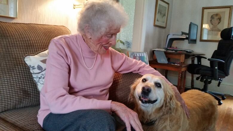 An elderly woman with white hair sits on a couch while petting a golden retriever.