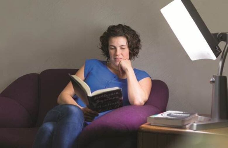 A woman sits in an armchair reading a book. A light therapy lamp is on next to her. 