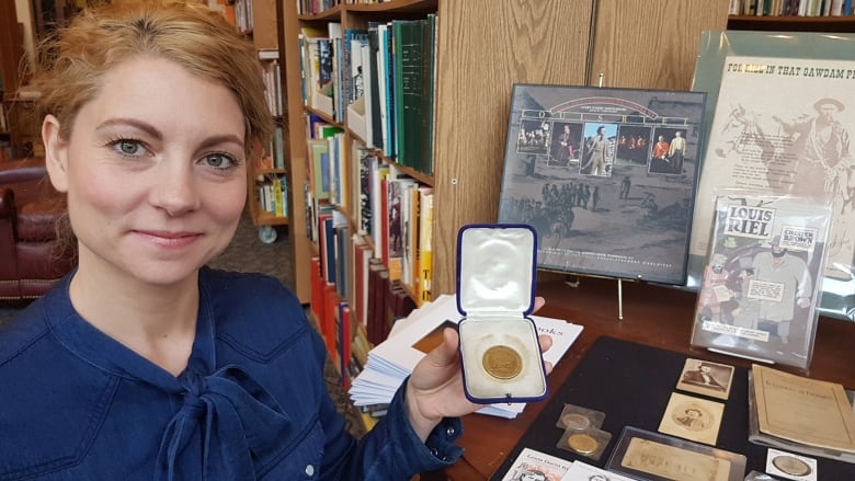 A woman holds up a small box with a medallion as she stands in front of shelves of books.