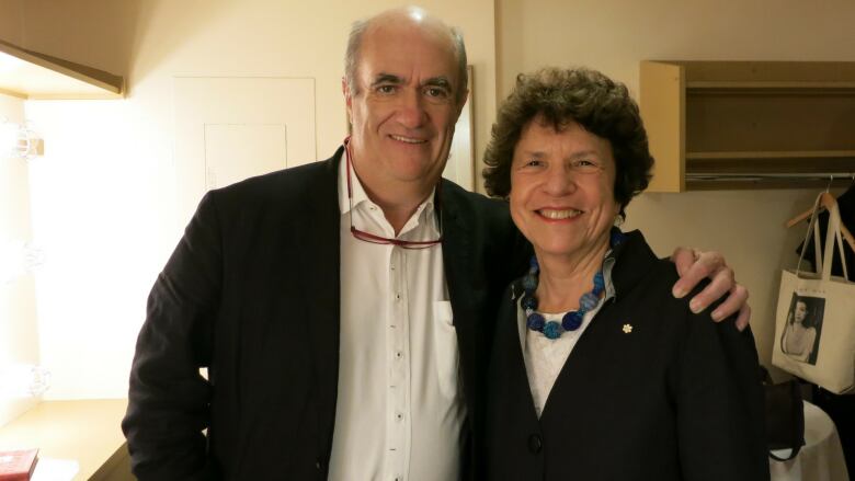 A man in a suit smiles at the camera with his arm around a woman with curly short hair.