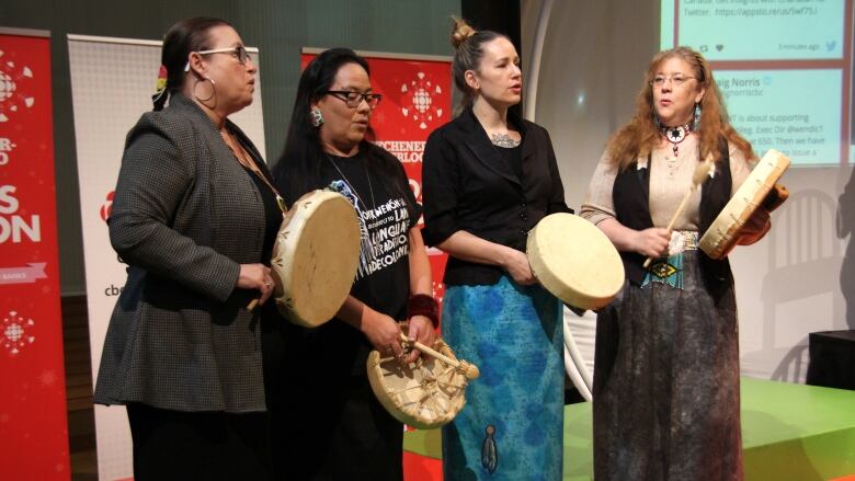 Four individuals with Indigenous drums stand on a stage and sing