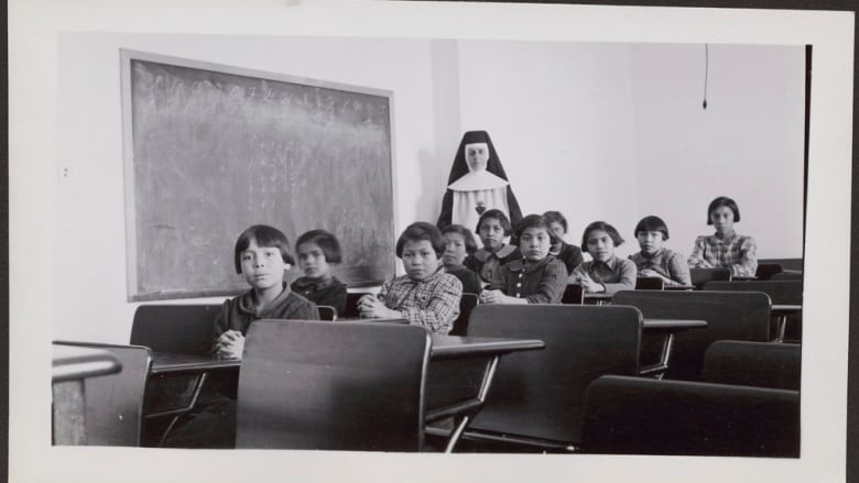 A black and white photo shows young students sitting in desks looking at the camera, with a woman in a nun's habit standing behind them.