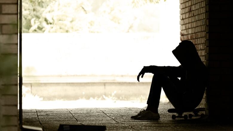 A person wearing a hoodie, sitting on a skateboard and leaning against a brick wall, is seen in silhouette.