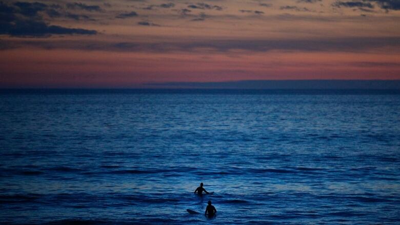 The silhouette of two surfers in deep rich blue waters are in front of a gorgeous sunset with hues of magenta, orange and peach.