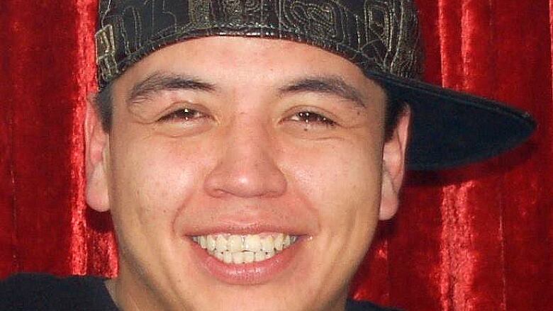 A youthful-looking Indigenous man with a sideways baseball cap smiles wide at the camera in front of a red velour background.