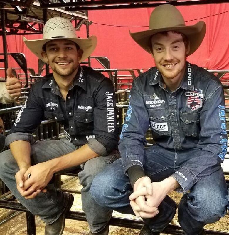 Two men wearing denim and cowboy hats sit on a gate