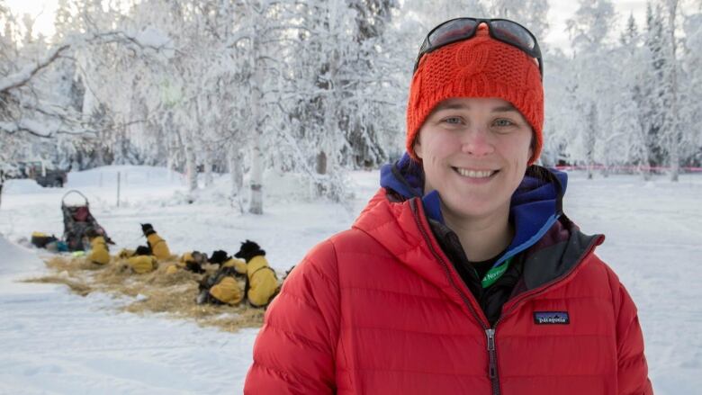 A woman in a red jacket and toque stands in front of sled dogs, lying on hay. 