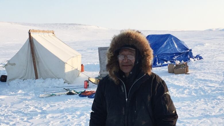 A man in a parka stands outside at a winter campsite.