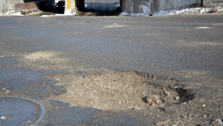 A large pothole is in the middle of College Street in Sudbury heading down towards a railroad underpass with crumbling concrete. 