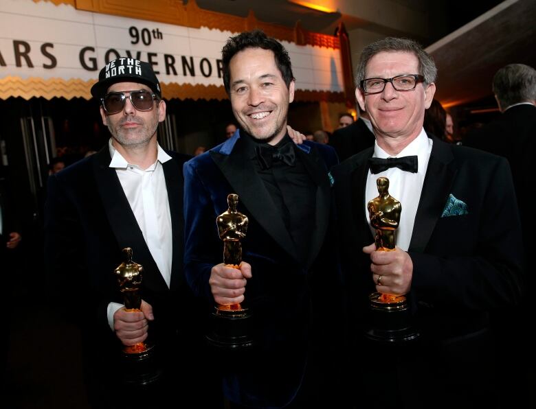 Three men in tuxedos hold Oscar statuettes. 