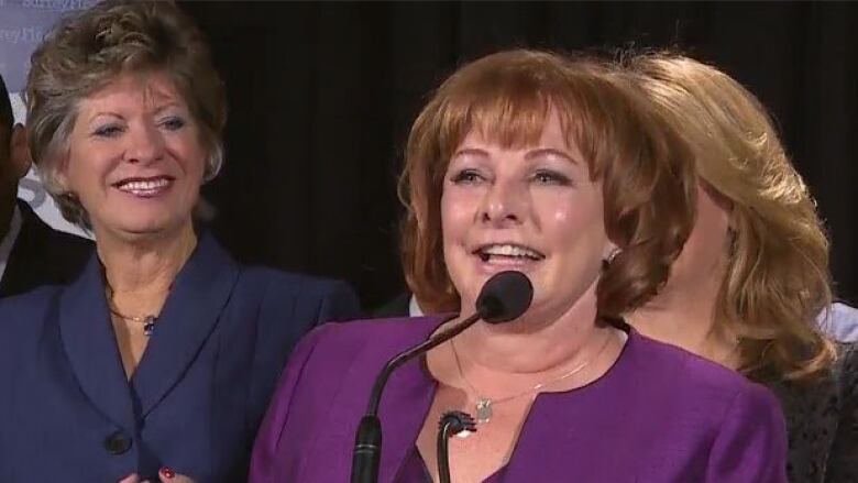 white woman with reddish hair and purple dress at microphone. two other white women in background