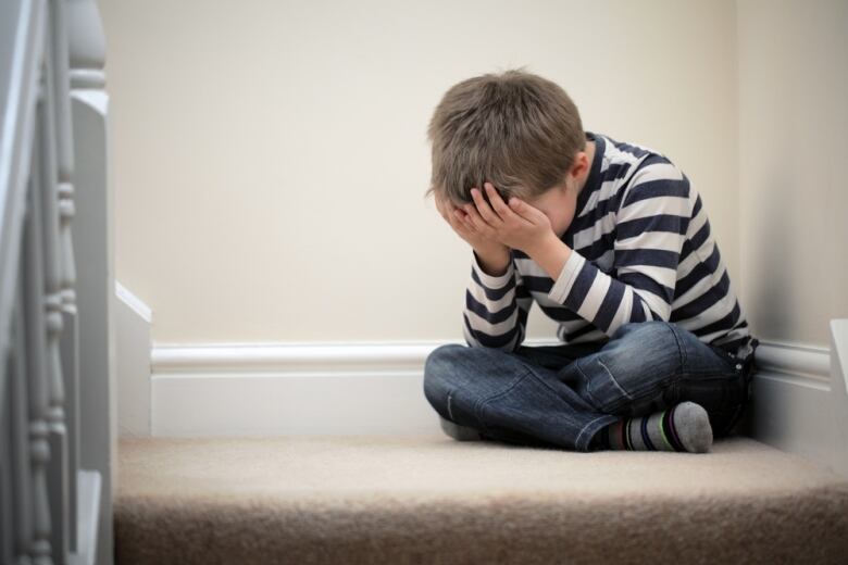 A boy sits in a corner and holds his head in his hands.