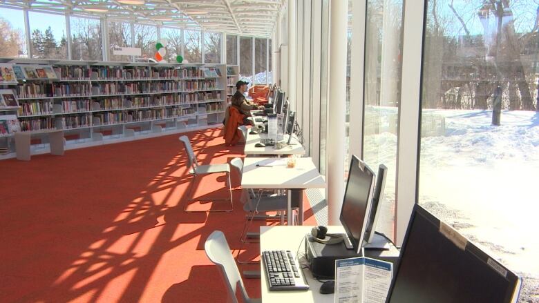 A row of tables with computers on them are set up in front of large windows. Rows of books can be seen in the back ground. 