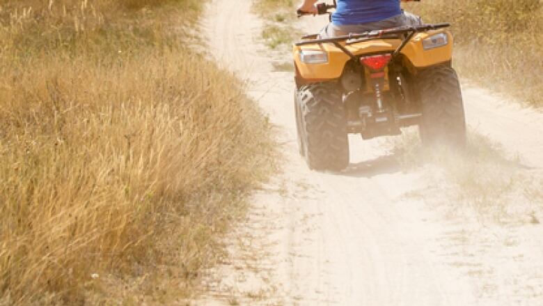 A person wearing a blue T-shirt drives a yellow ATV down a gravel roadway surrounded by long grass.