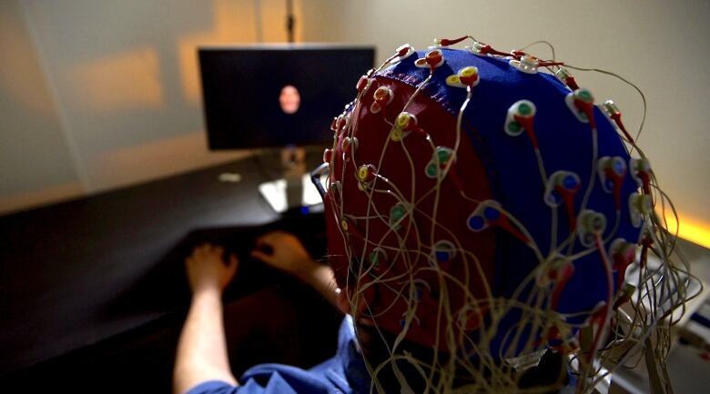 A person wears a cap on their head with sensor nodes and wires while watching a display monitor.