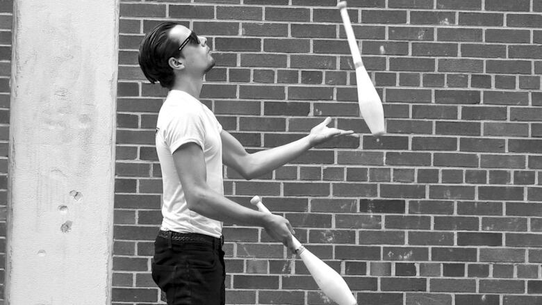 Man with slicked back hair and a white t-shirt juggles bowling pins in front of a brick wall.