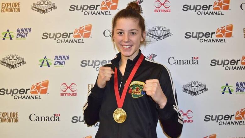 A young woman stands with her arms up, read to box, wearing a gold medal.