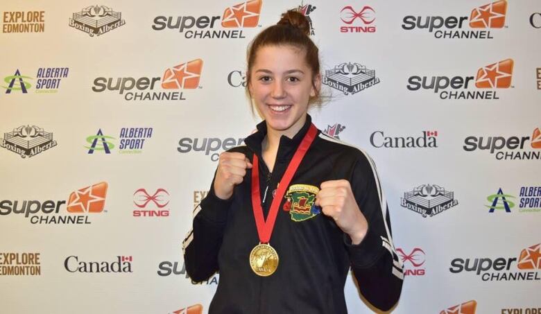 A young woman stands with her arms up, read to box, wearing a gold medal.