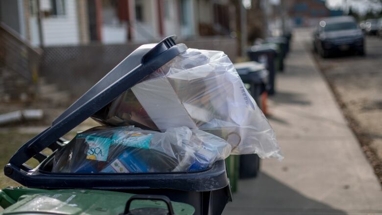 A recycle bin on a sidewalk overflows with items contained in a plastic bag.