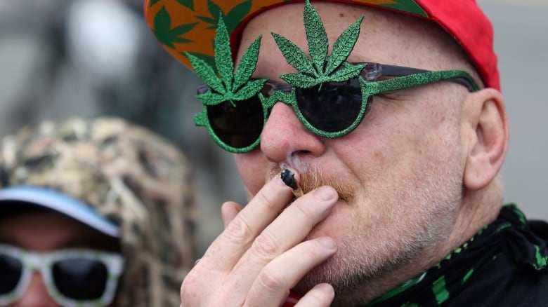 A man with white stubble on his face wearing a red ball cap smokes a joint while wearing green glasses with little pot leafs on the rim