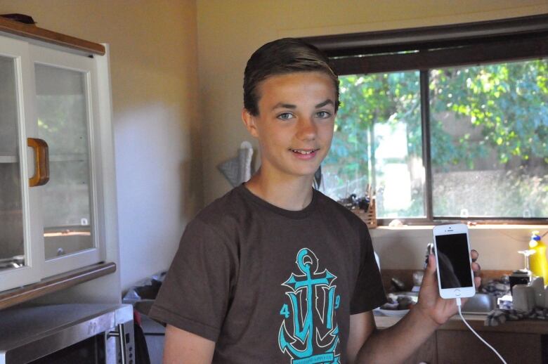 A young boy, his hair neatly combed to the side and wearing a brown t-shirt, holds up his iPhone while grinning at the camera, light from the window behind him filtering into the room.