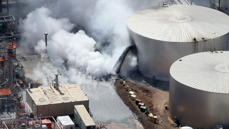 Thick white smoke billows as a black liquid pours out of a large refinery tank.