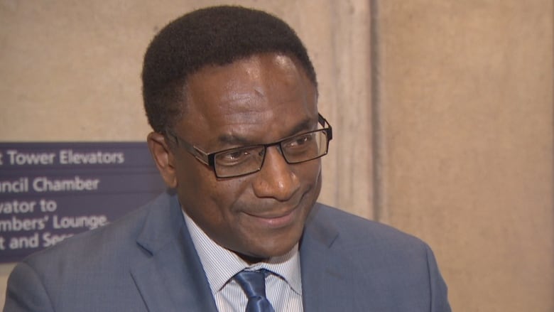 A head and shoulders shot of a middle aged Black man in a suit and tie standing inside a civic building with wood panelling. A microphone is visible at the bottom of the frame