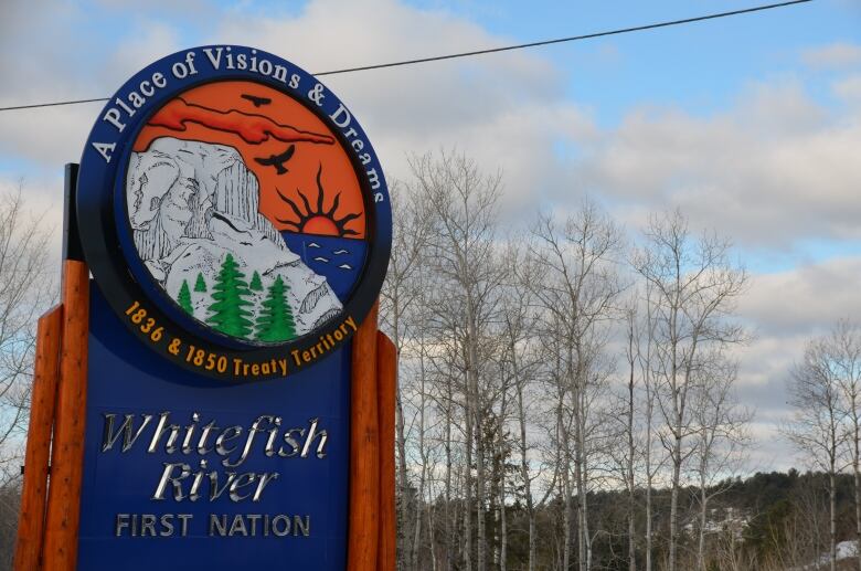 A blue and orange sign depicting a mountain, forest and rising sun welcomes people to the Whitefish River First Nation