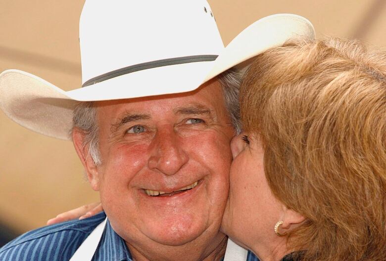 Premier Ralph Klein is smiling in a cowboy hat as a woman gives him a kiss on the cheek.