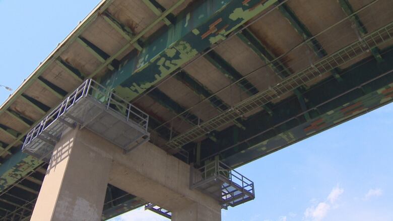 Spotty paint and a metal walkway suspended under a bridge.