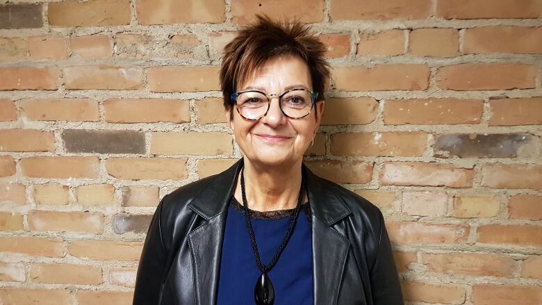 A woman with short brown hair in a leather jacket stands against a brick wall.