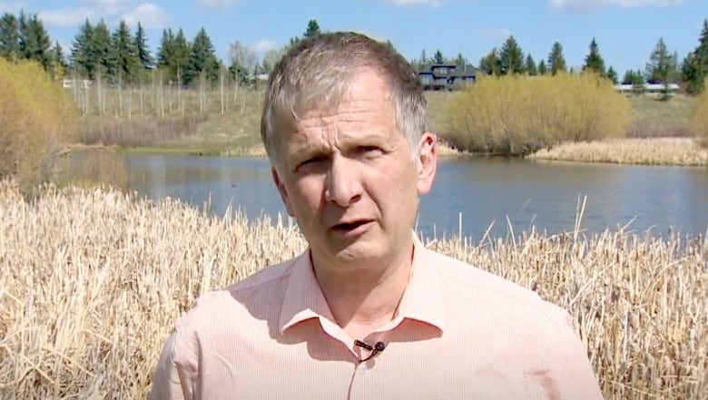 A man in a light shirt is seen standing in daylight with a body of water and shrubs in the background.