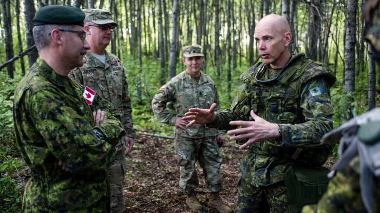 Canadian soldiers in green camo stand talking in a circle in a wooded area.