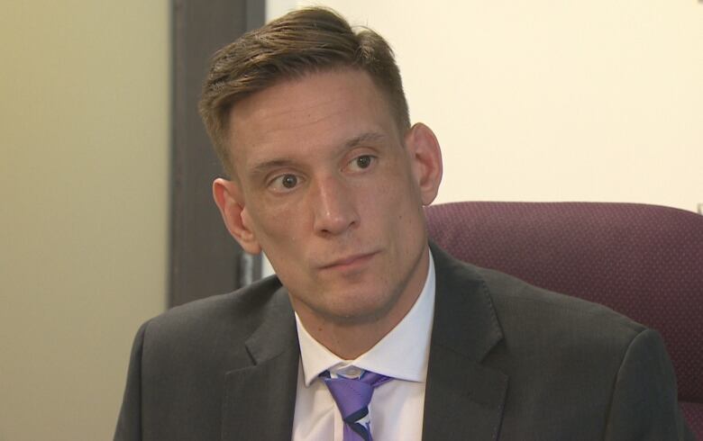 A man with short hair and a grey suit, white shirt and purple tie looks away.