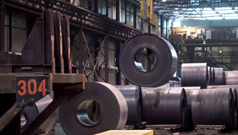 Large steel drums are seen on a factory floor, labelled with numbers and being lifted by a crane.