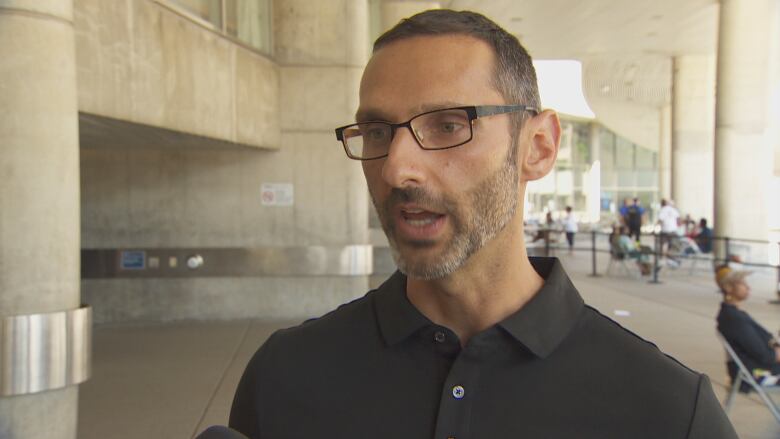 A man with glasses and a close-shaved beard does an interview in a building with people sitting in the background.