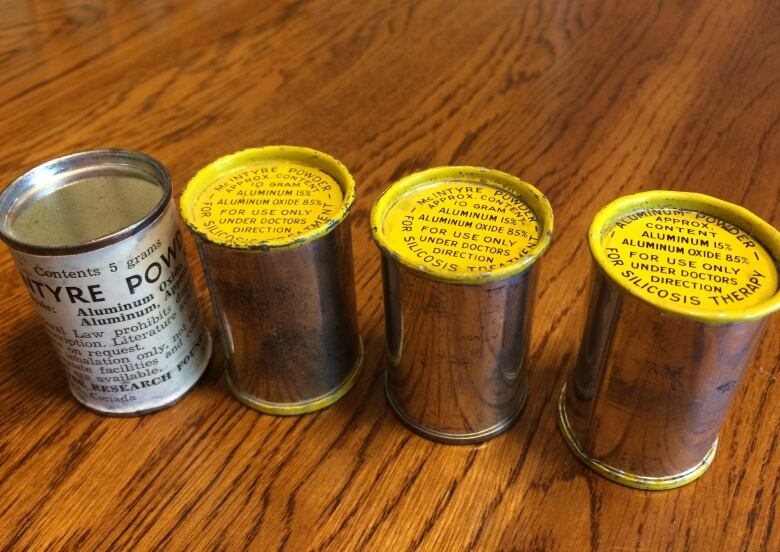 Four small canisters, one white with black writing, the other three shiny with yellow tops and bottoms with black writing, sitting on a woodgrain table.