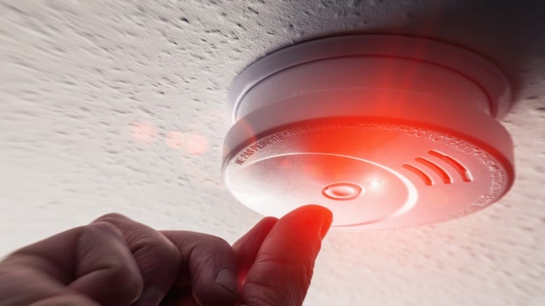 A smoke detector on a ceiling with the red light flashing and a hand reaching up towards it. 