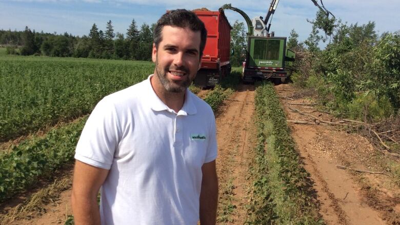 Man with brown hair and a beard, wearing a white polo shirt, stands in a field where wood debris is being turned into wood chips. 