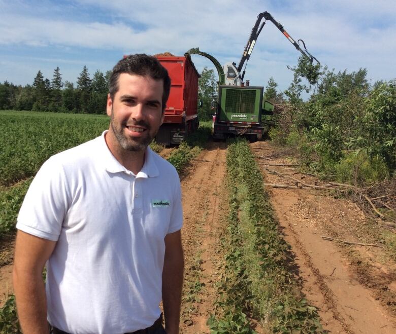 Man with brown hair and a beard, wearing a white polo shirt, stands in a field where wood debris is being turned into wood chips. 