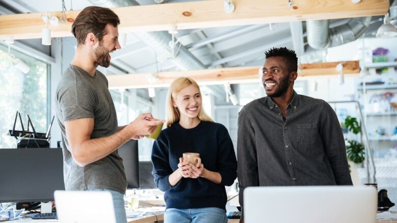 Three people laugh and smile together while holding mugs and standing in an office. 