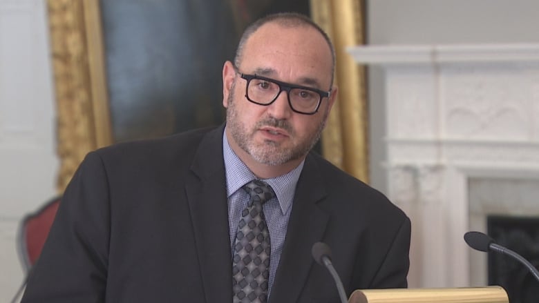 A man with short hair and short beard and moustache wearing dark-rimmed glasses, dark blue suit, light blue shirt and dark blue patterned tie speaks into a microphone.