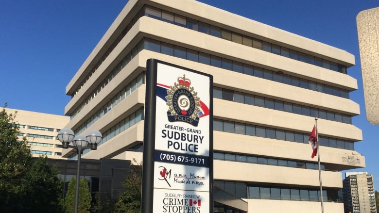 Th picture shows a seven-storey square building with a sign with the Greater Sudbury Police logo and phone number and a Canadian flag against a blue sky.