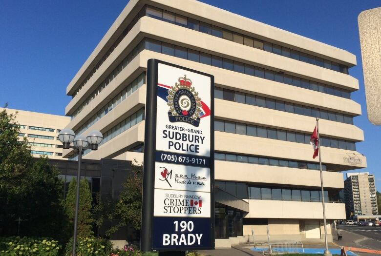 Th picture shows a seven-storey square building with a sign with the Greater Sudbury Police logo and phone number and a Canadian flag against a blue sky.